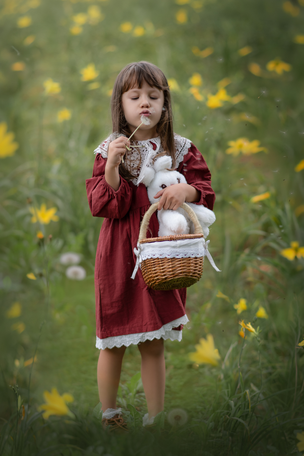 Uma criança usando um vestido vermelho entre flores amarelas, soprando um dente de leão durante o ensaio artistico infantil.