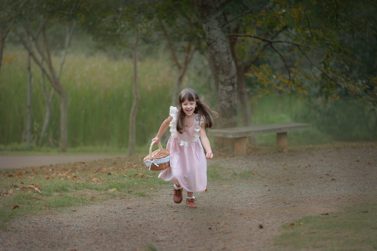 Uma crianças usando um vestido rosa, estilo camponesa e brincando em um parque com folhas, durante seu ensaio artistico.
