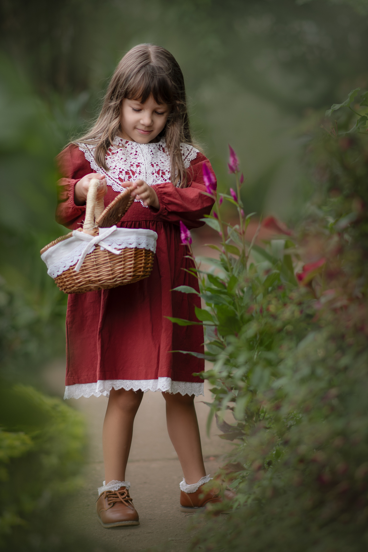 Uma criança usando vestido vermelho e colhendo flores com uma cesta vintage, durante ensaio artístico infantil na natureza.