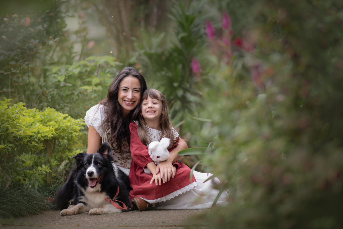 Uma crianças usando um vestido rosa, estilo camponesa junto com sua mãe e brincando com um coelho de pelúcia e sua cachorra Border Collie, em um parque, durante seu ensaio artistico.