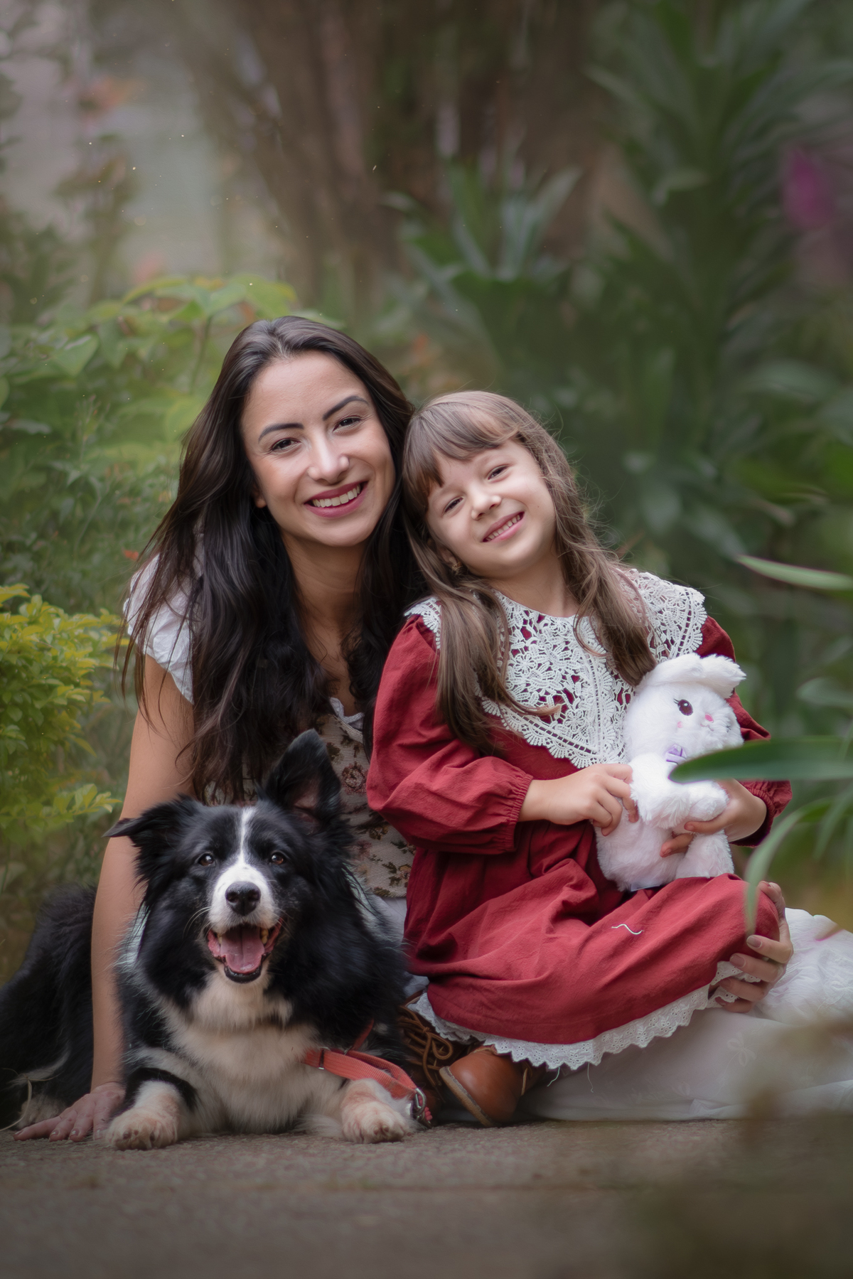 Uma crianças usando um vestido rosa, estilo camponesa junto com sua mãe e brincando com um coelho de pelúcia e sua cachorra Border Collie, em um parque, durante seu ensaio artistico.