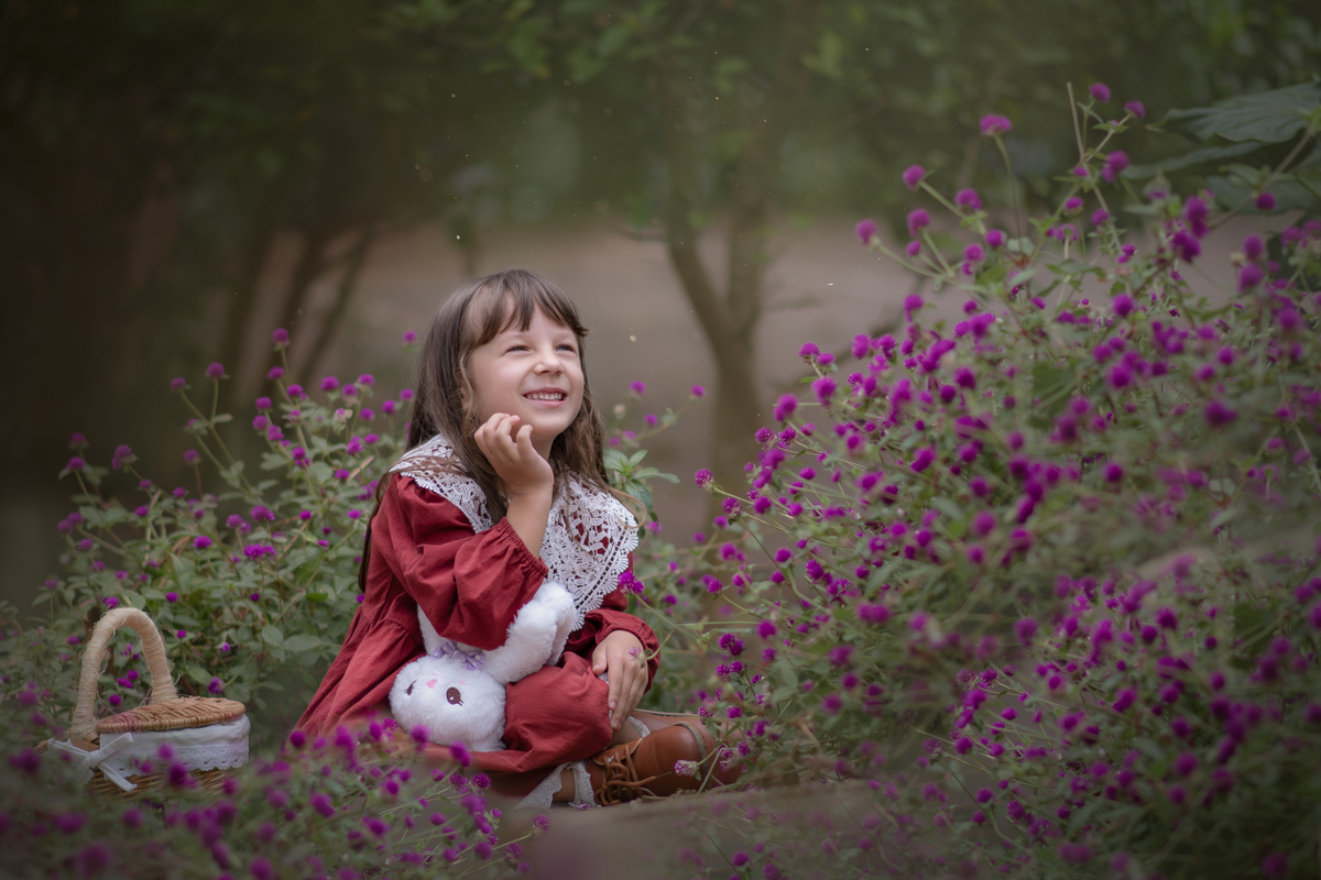 Uma criança usando um vestido vesmelho entre flores roxa, durante o ensaio fotográfico infantil externo.
