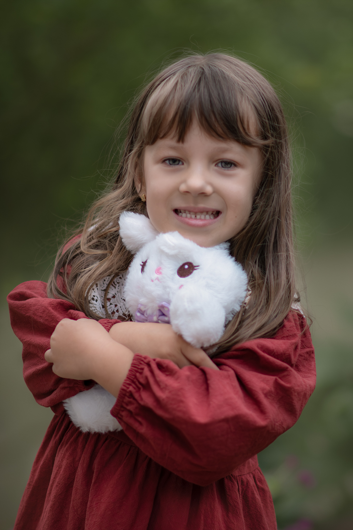 Uma criança usando vestido vermelho e abraçando um coelho de pelúcia branco, durante seu ensaio artístico infantil externo.