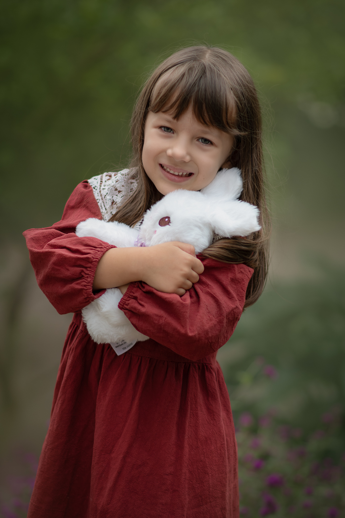 Uma criança usando vestido vermelho e abraçando um coelho de pelúcia branco, durante seu ensaio artístico infantil externo.