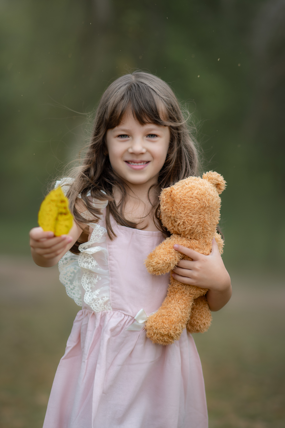Uma crianças usando um vestido rosa bem estilo camponesa e brincando com um ursinho de pelúcia marrom, em um parque, durante seu ensaio artistico.