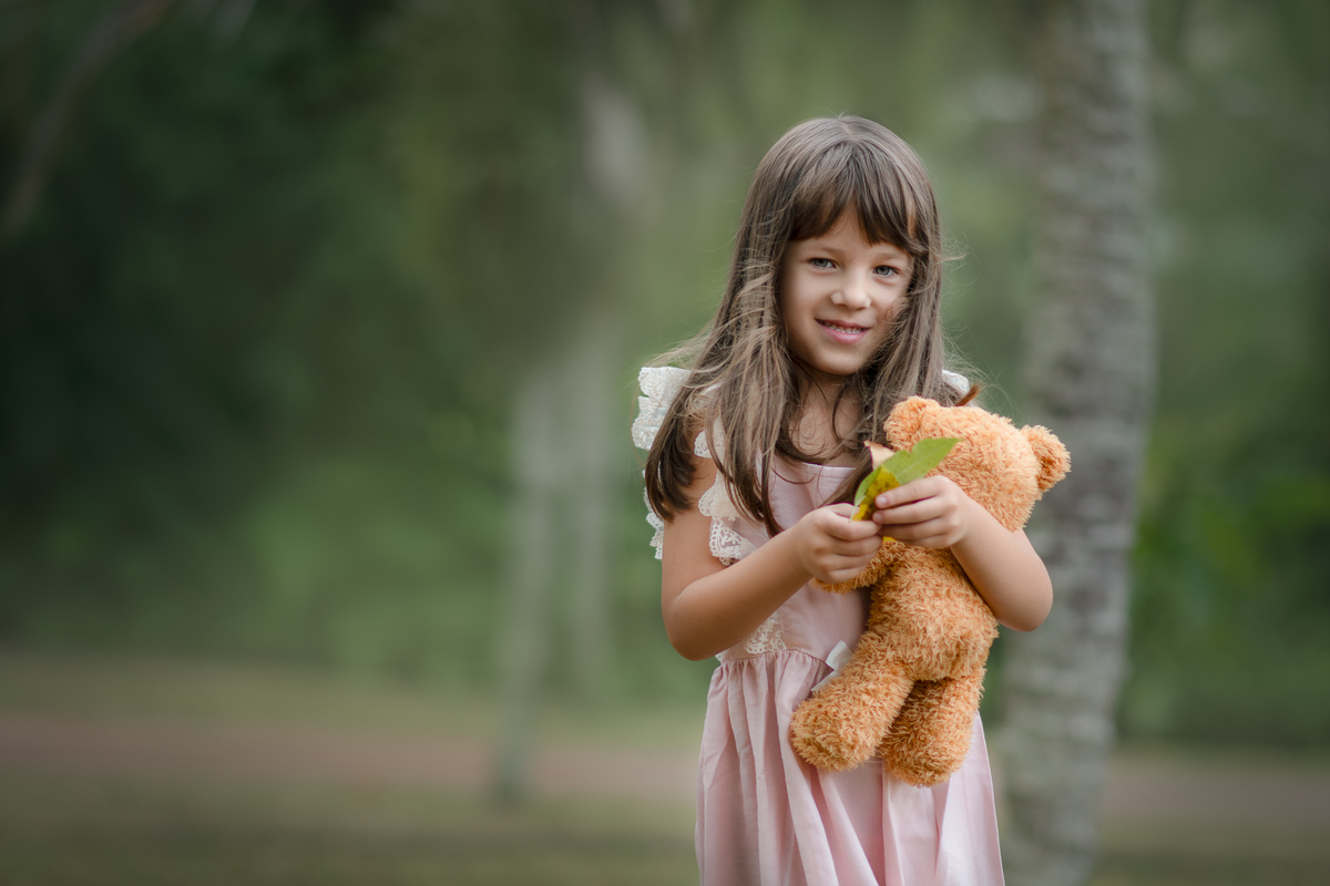 Uma crianças usando um vestido rosa bem estilo camponesa e brincando com um ursinho de pelúcia marrom, em um parque, durante seu ensaio artistico.