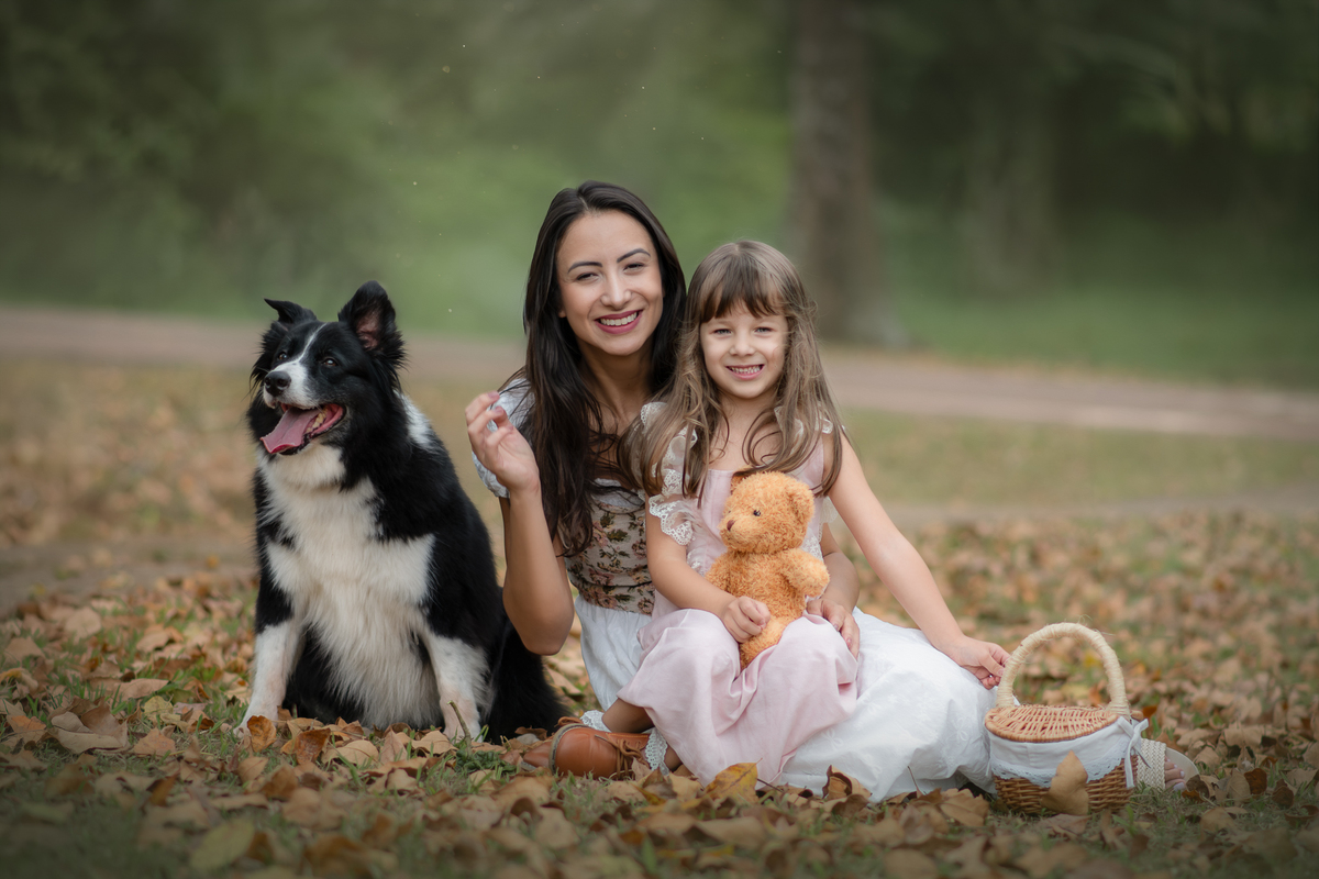 Uma crianças usando um vestido rosa, estilo camponesa junto com sua mãe e brincando com um ursinho de pelúcia marrom e sua cachorra Border Collie, em um parque, durante seu ensaio artistico.