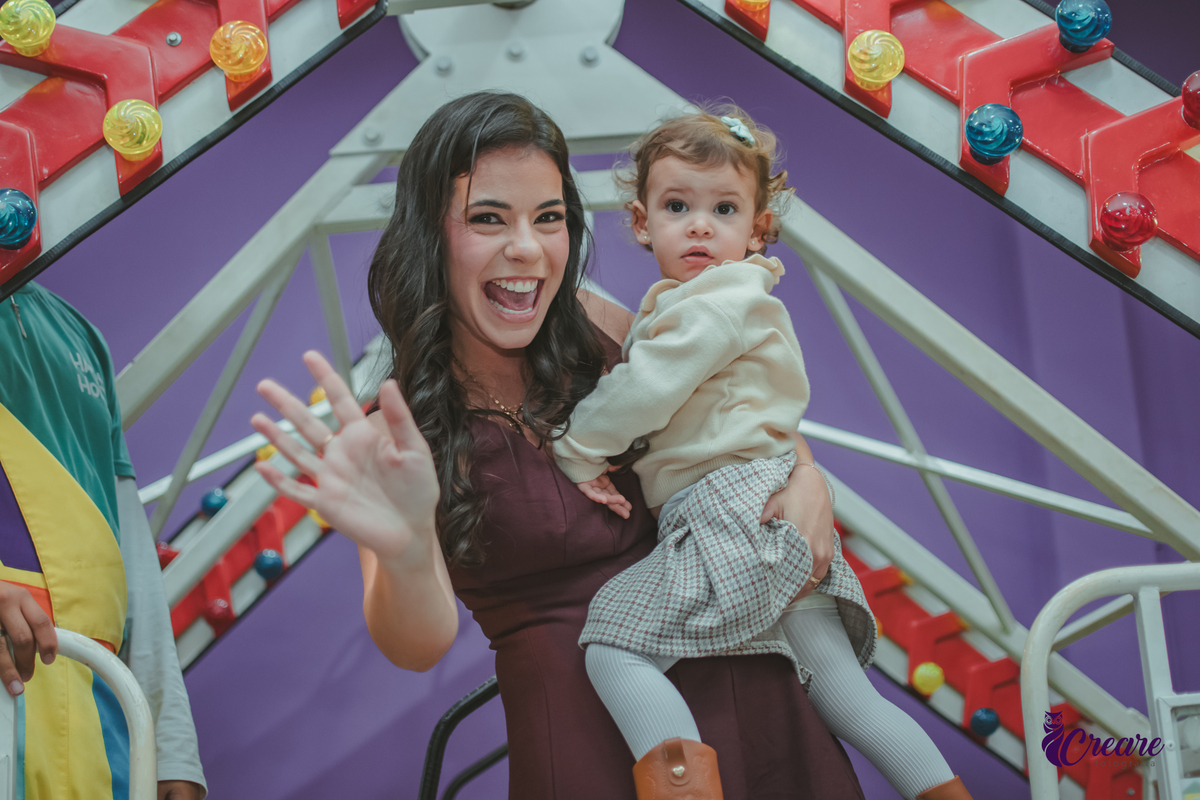 Uma mulher com uma menina de 2 anos no colo, dando tchau ao sair do brinquedo Barco viking durante festa de aniversário infantil.
