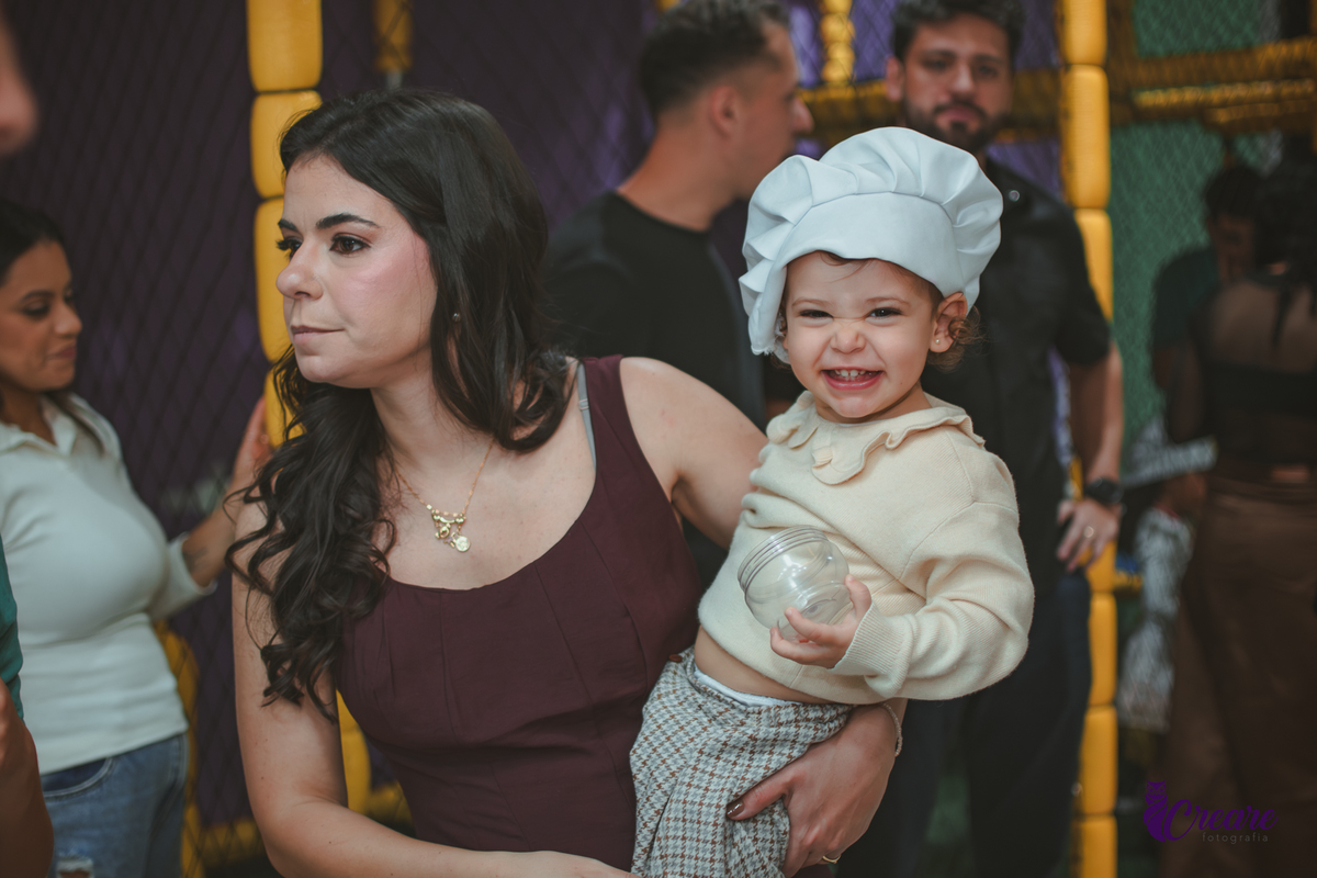 Uma criança de 2 anos usando uma touquinha e sorrindo no colo da sua mãe, durante sua festa de aniversáio no Buffet Happy House em Santo André.