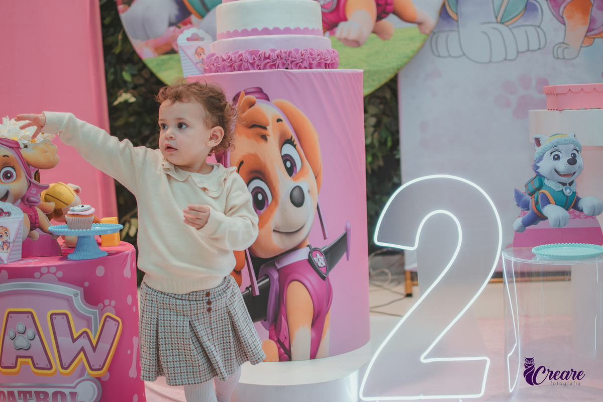 Uma criança de 2 anos em frente a mesa de decoração do seu anversário com tema patrulha canina rosa, ao lado da criança um letreiro brilhante de número 2.