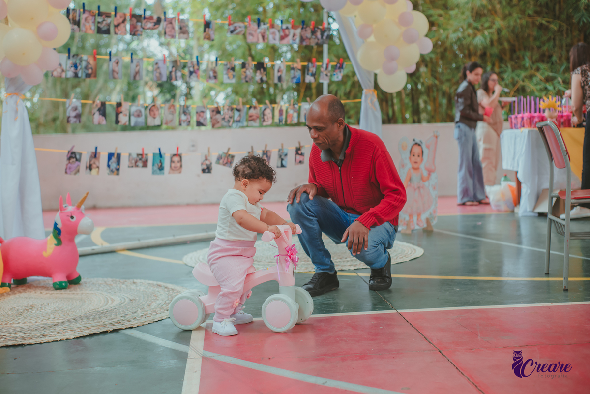 Uma homem negro, ajudando uma bebê andar de bicicletinha durante festa de aniversário de 1 ano.