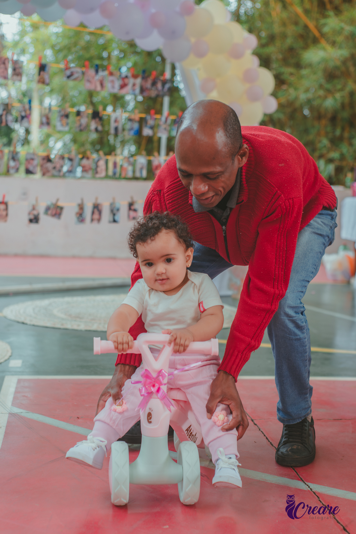 Uma homem negro, ajudando uma bebê andar de bicicletinha durante festa de aniversário de 1 ano.