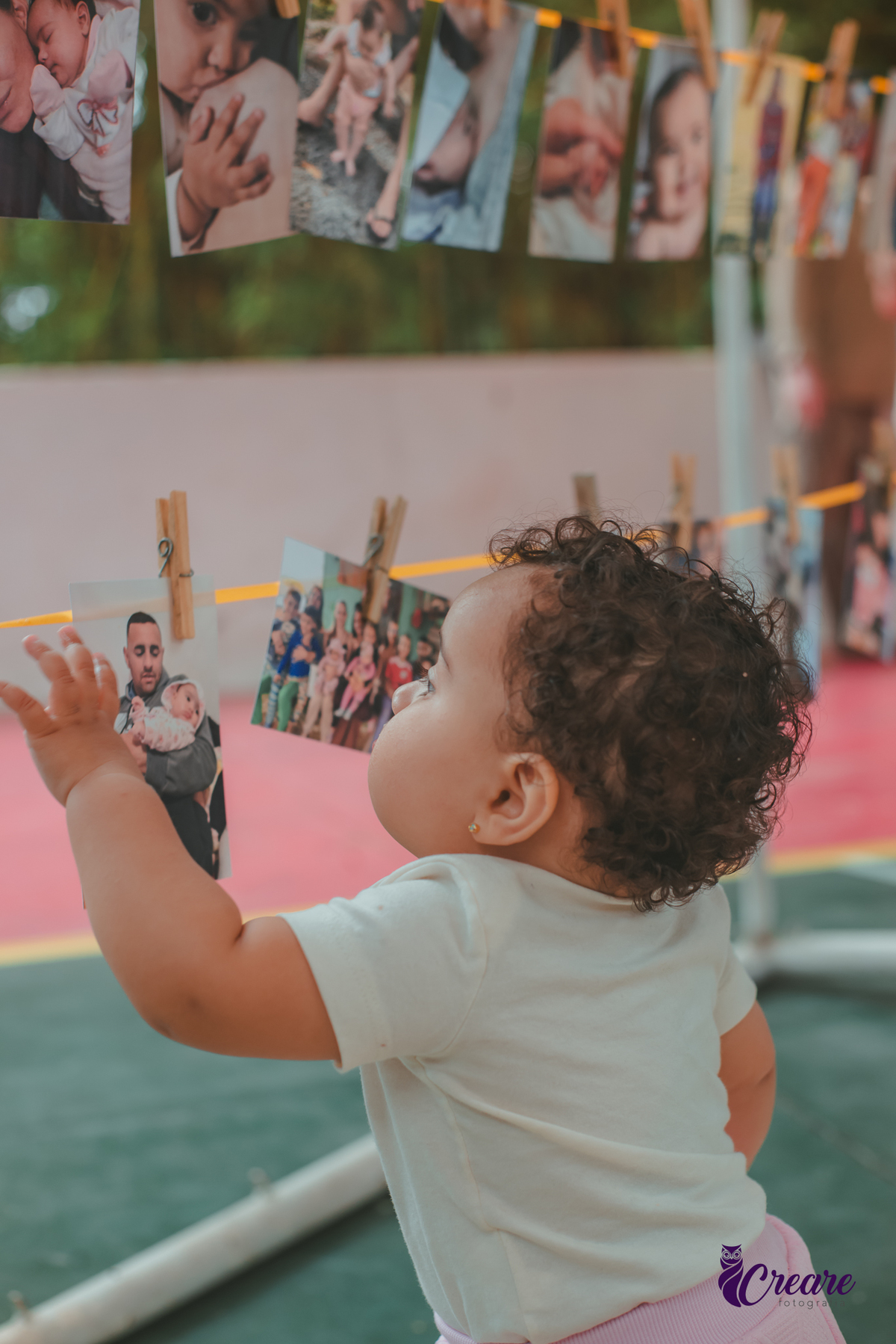 Uma bebê observando algumas fotografias pensurdas em um varal de fotos, durante festa de aniversário de 1 ano.