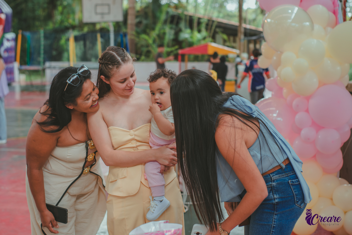 Varias mulheres observando uma bebê e sorrindo, durante festa de aniversário de 1 ano.
