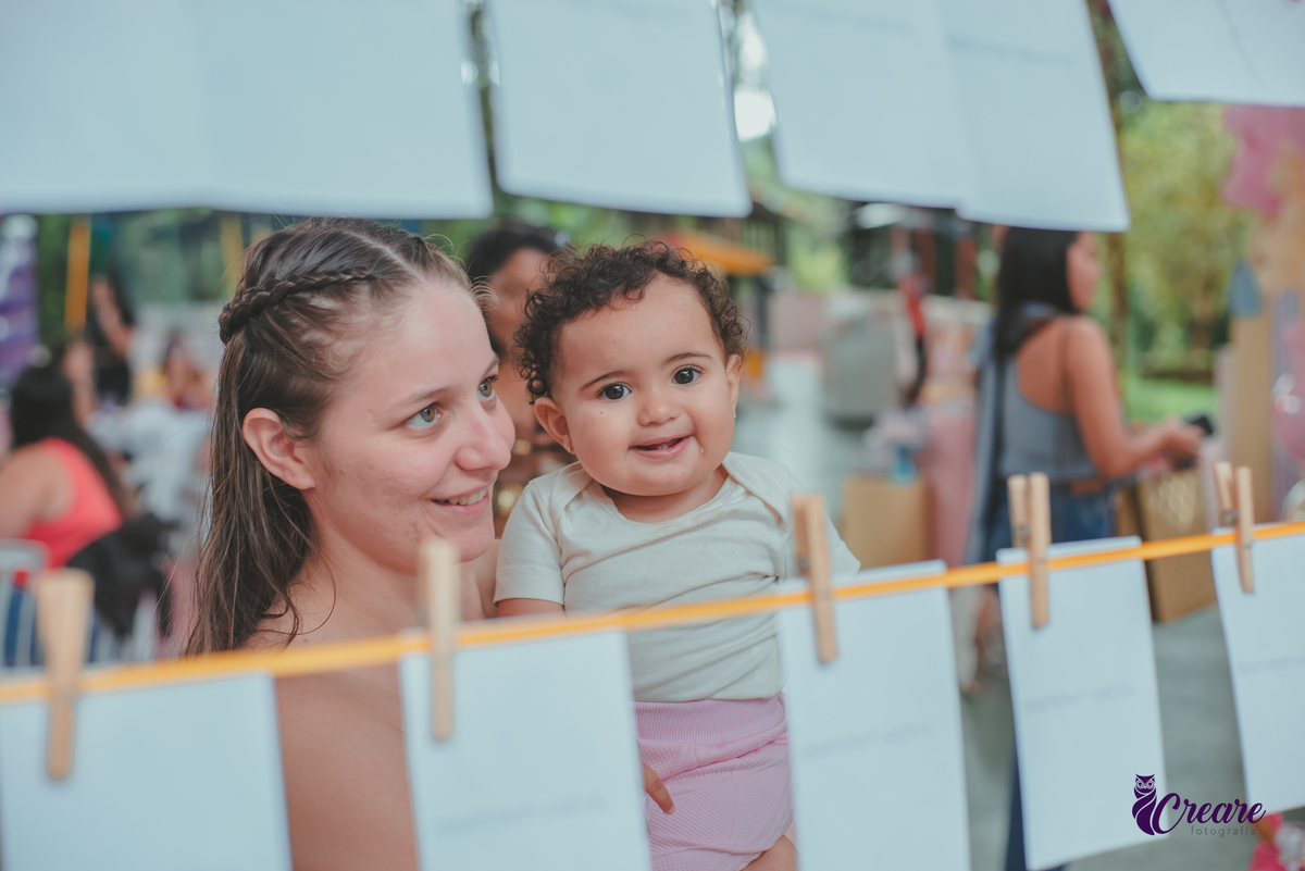 Uma mulher e uma bebê observando algumas fotografias pensurdas em um varal de fotos, durante festa de aniversário de 1 ano.