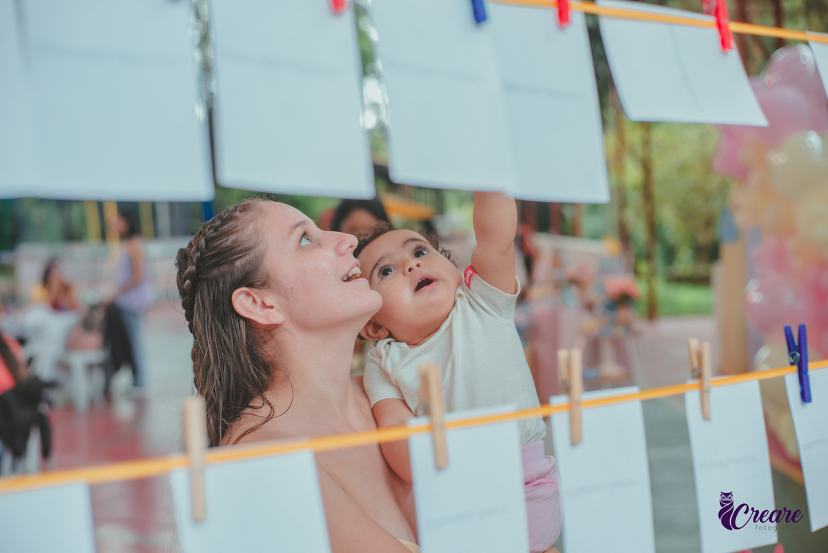 Uma mulher e uma bebê observando algumas fotografias pensurdas em um varal de fotos, durante festa de aniversário de 1 ano.