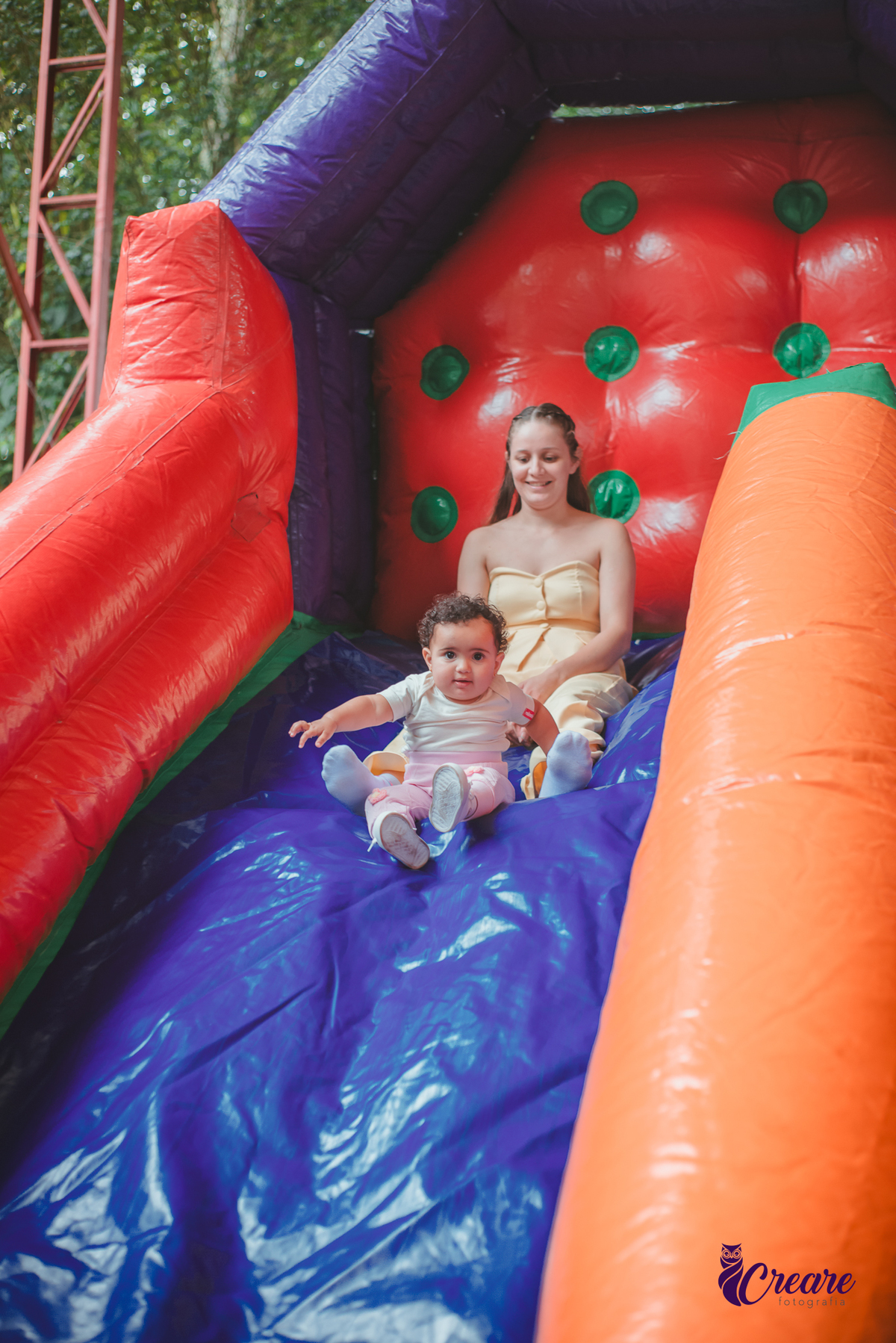 Uma mulher brincando no escorregador de ar, com sua bebê de 1 ano, durante festa de aniversário.