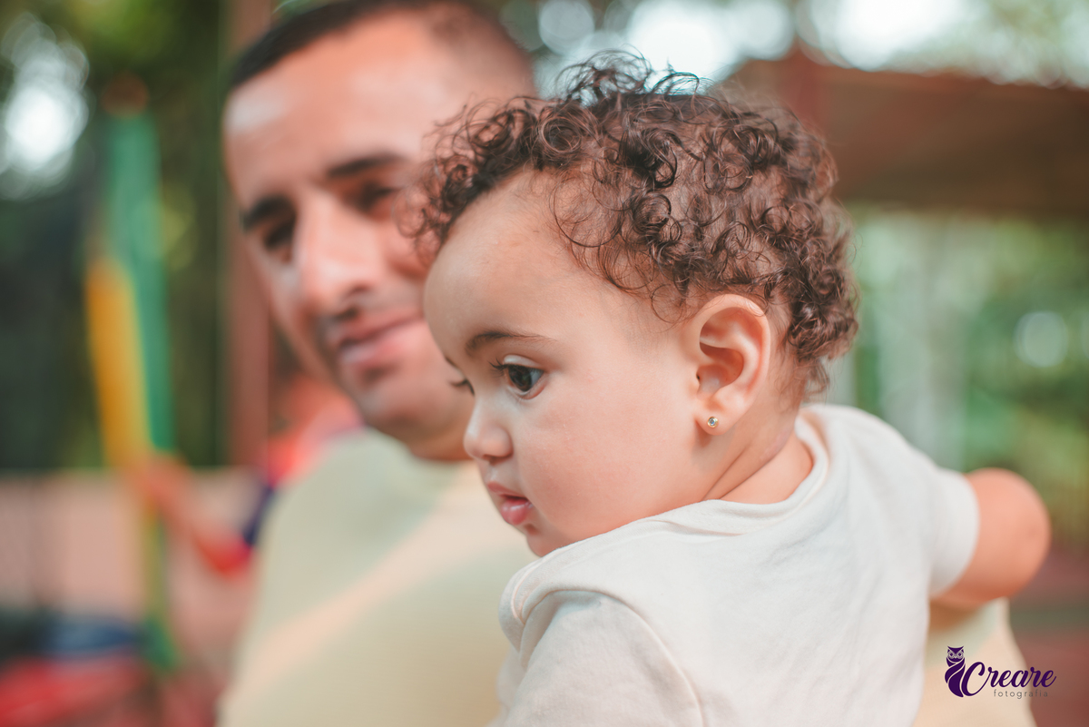 Uma homem com uam bebê de colo em primeiro plano, durante festa de aniversário de 1 ano.