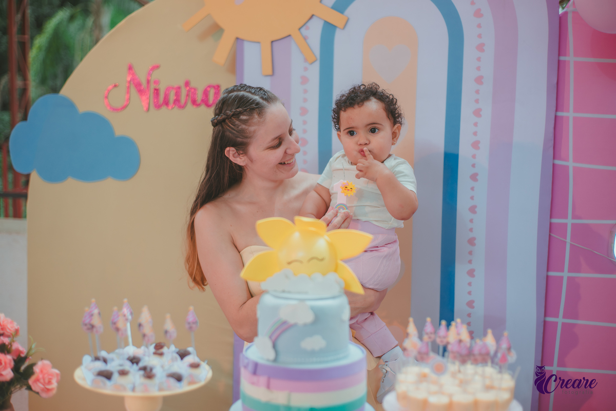 Uma mãe com sua bebê em frente uma mesa de decoração com tema Primeira volta ao sol, durante festa de aniversário.