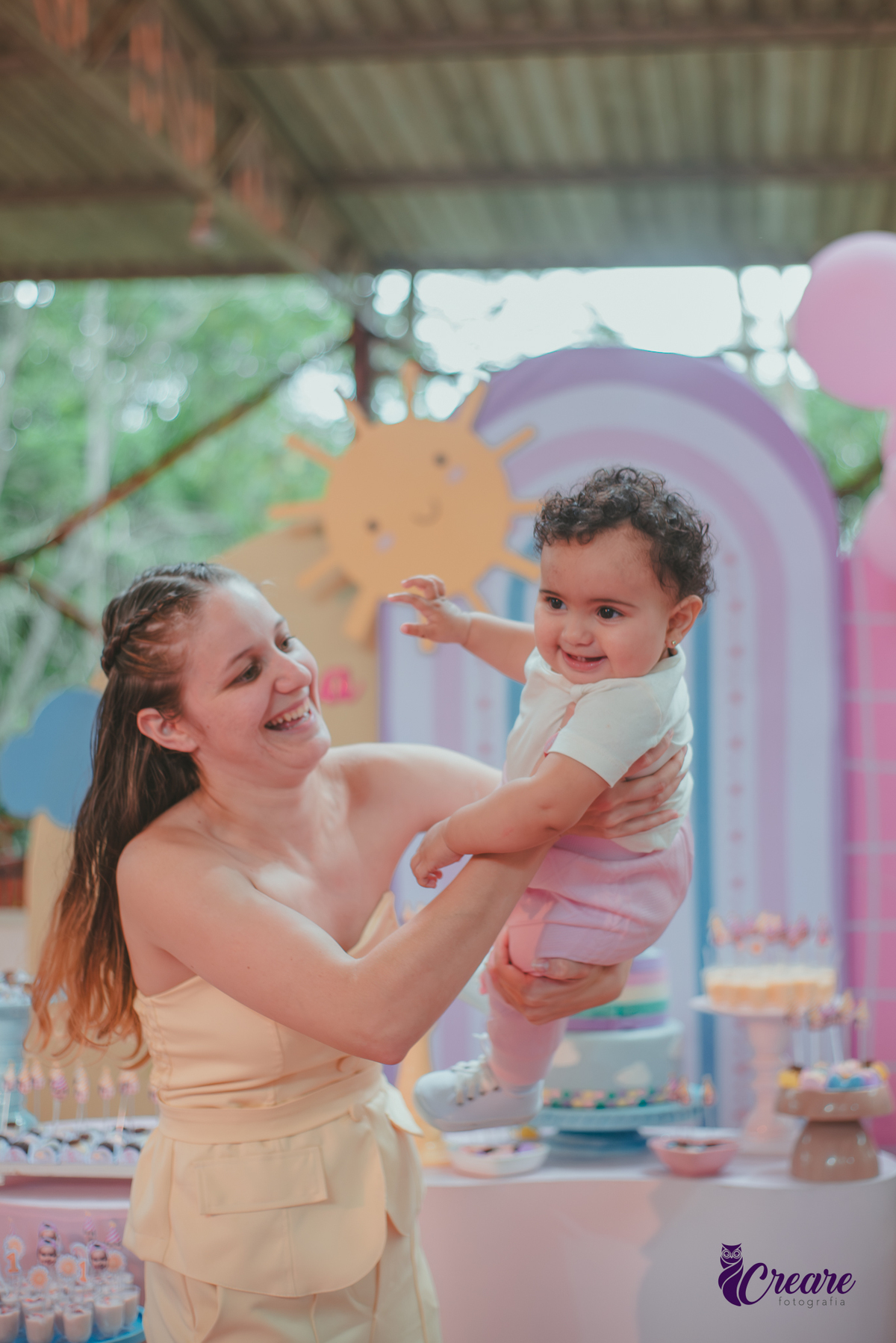 Uma mãe brincando com sua bebê em frente uma mesa de decoração com tema Primeira volta ao sol, durante festa de aniversário.
