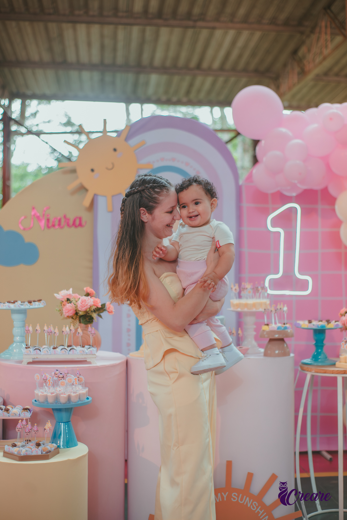 Uma mulher com roupa amarela, brincando com sua bebê em frente uma mesa de decoração com tema Primeira volta ao sol, durante festa de aniversário.