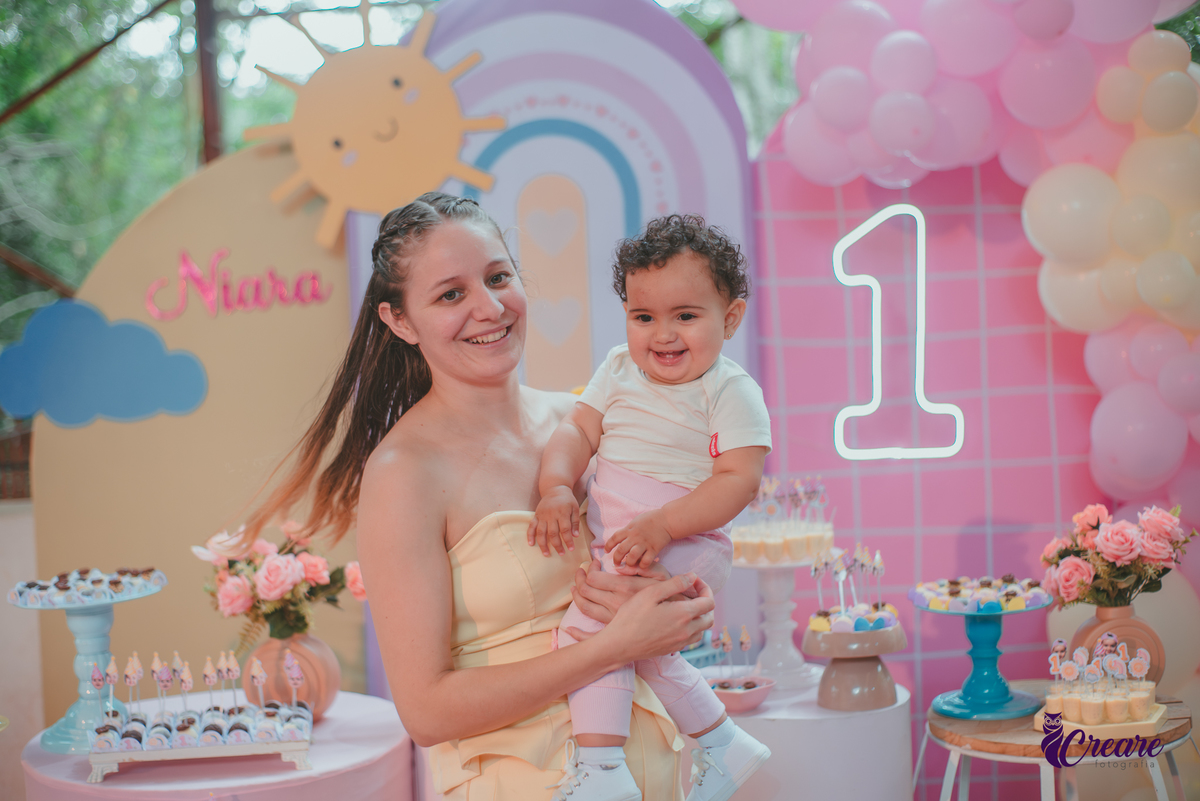 Uma mãe brincando com sua bebê em frente uma mesa de decoração com tema Primeira volta ao sol, durante festa de aniversário.
