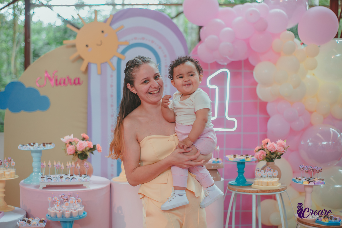 Uma mãe brincando com sua bebê em frente uma mesa de decoração com tema Primeira volta ao sol, durante festa de aniversário.