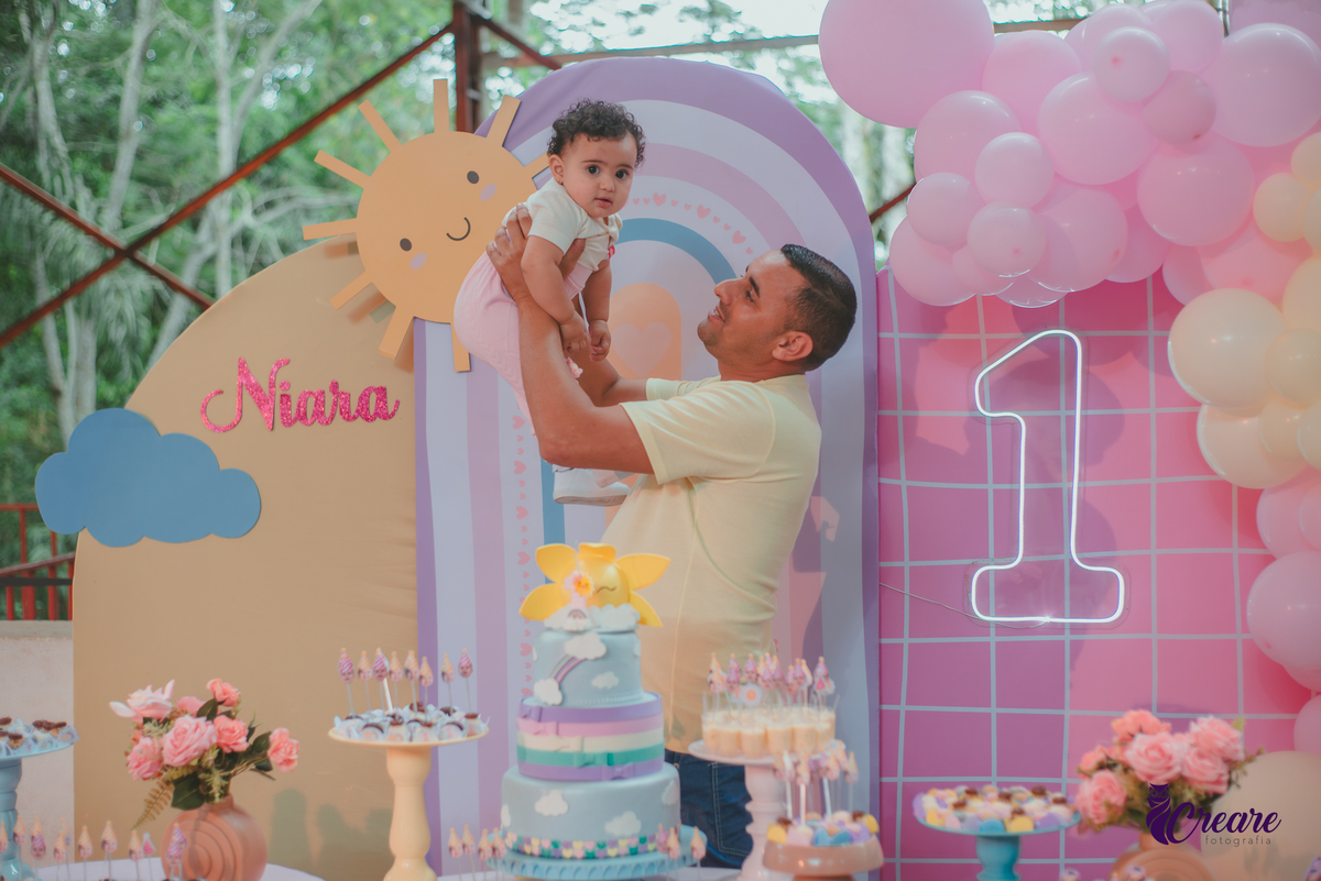 Um homem brincando com sua bebê em frente uma mesa de decoração com tema Primeira volta ao sol, durante festa de aniversário.