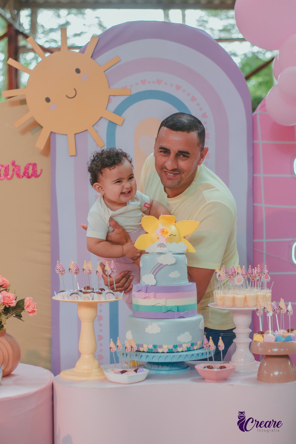 Um homem brincando com sua bebê em frente uma mesa de decoração com tema Primeira volta ao sol, durante festa de aniversário.
