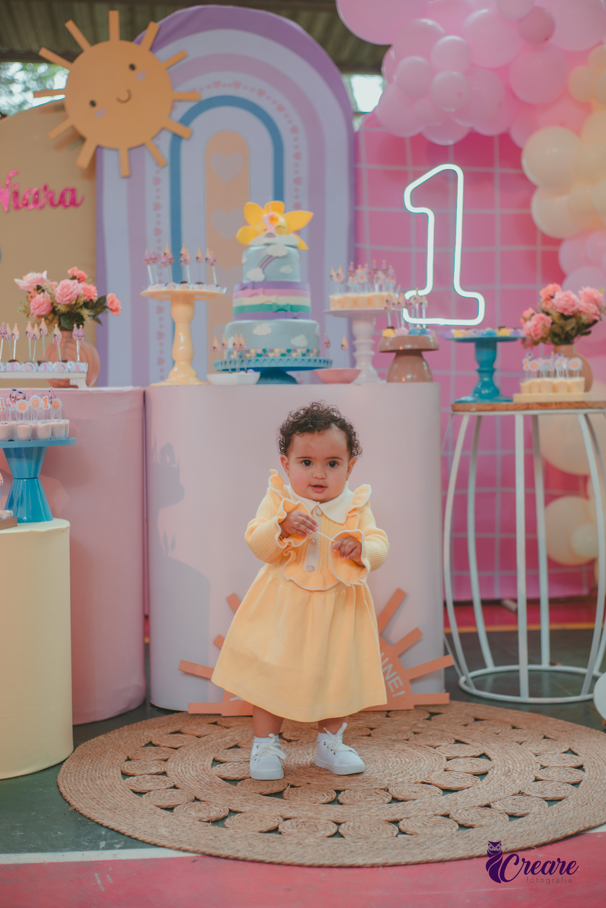 Uma bebê usando vestido amarelo e em frente mesa de decoração do seu aniversário de 1 ano.