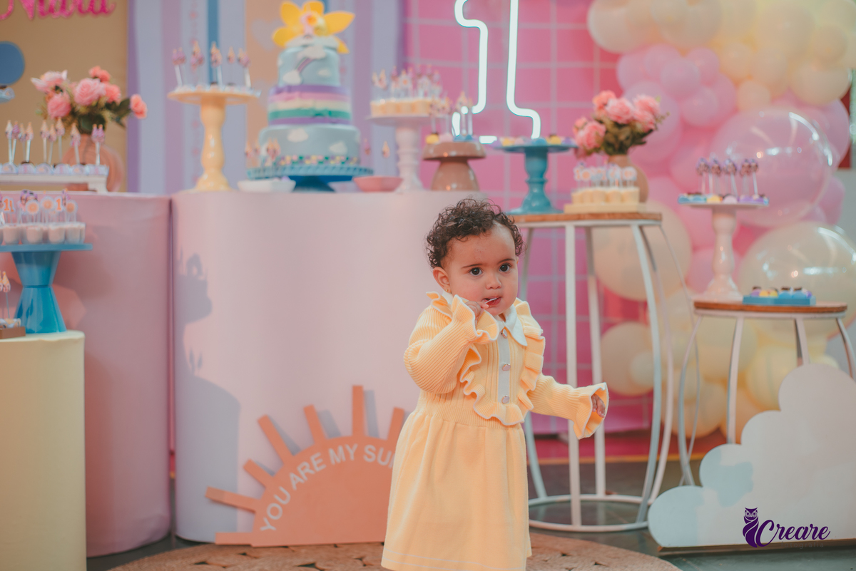 Uma bebê usando vestido amarelo e em frente mesa de decoração do seu aniversário de 1 ano.