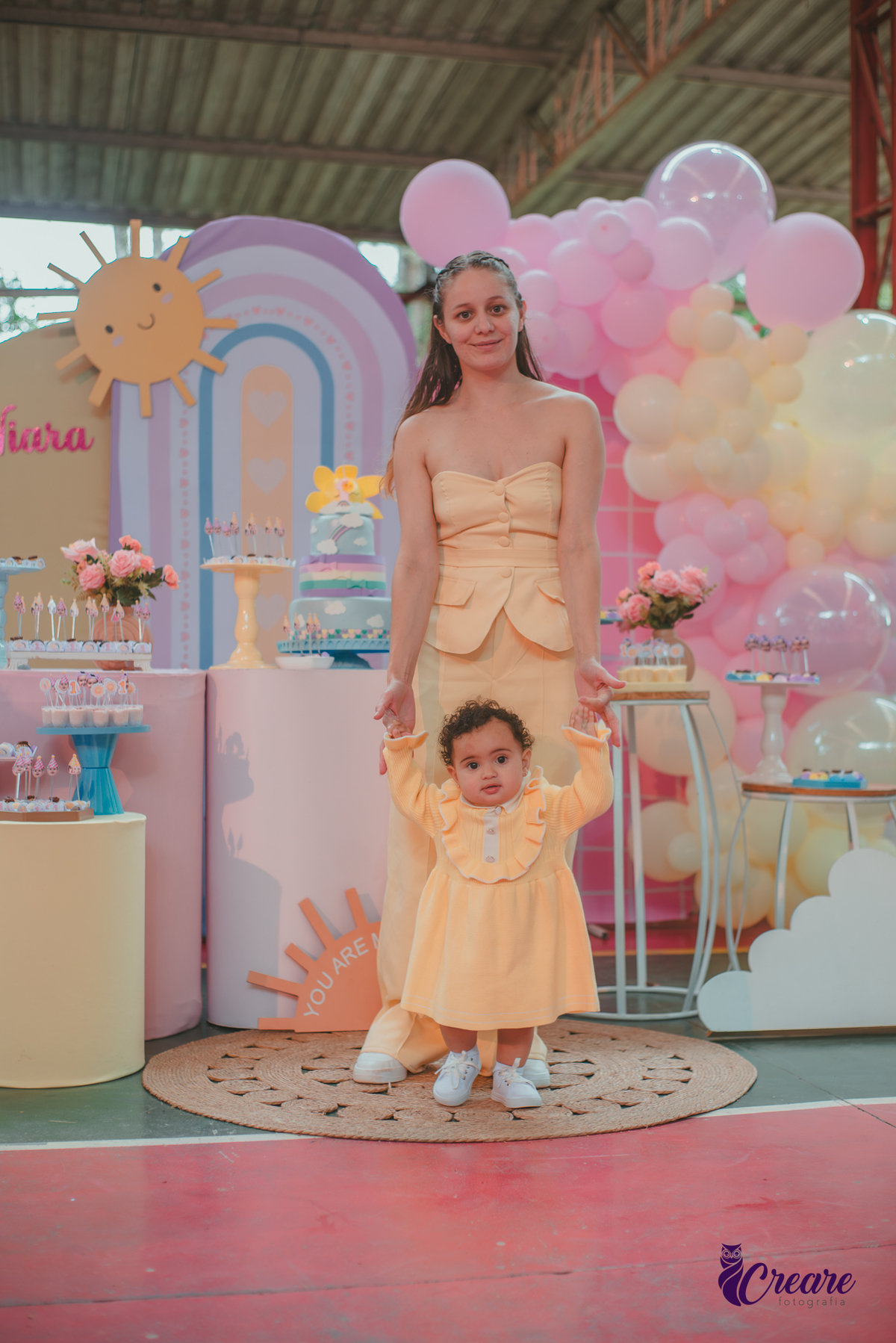 Uma bebê, usando vestido amarelo, e sua mãe em frente mesa de decoração do seu aniversário de 1 ano.