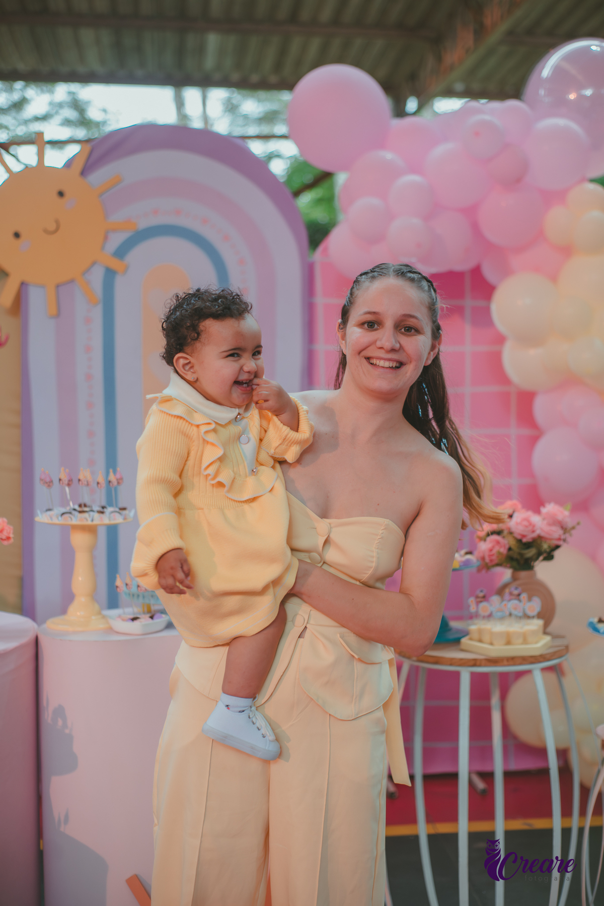 Uma mulher usando roupa amarela e uma bebê usando vestudo amarelo, sorrindo em frente a mesa de decoração do aniversário de 1 ano