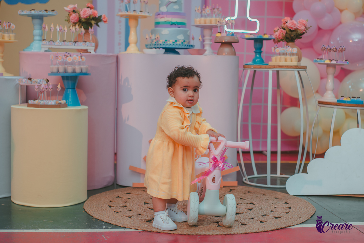 Uma bebê usando vestido amarelo e em frente mesa de decoração do seu aniversário de 1 ano.