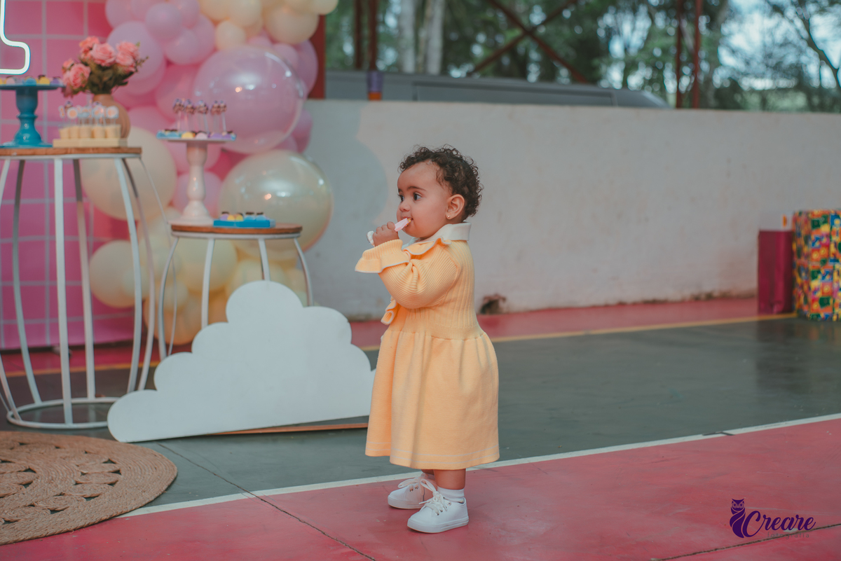 Uma bebê usando vestido amarelo e em frente mesa de decoração do seu aniversário de 1 ano.