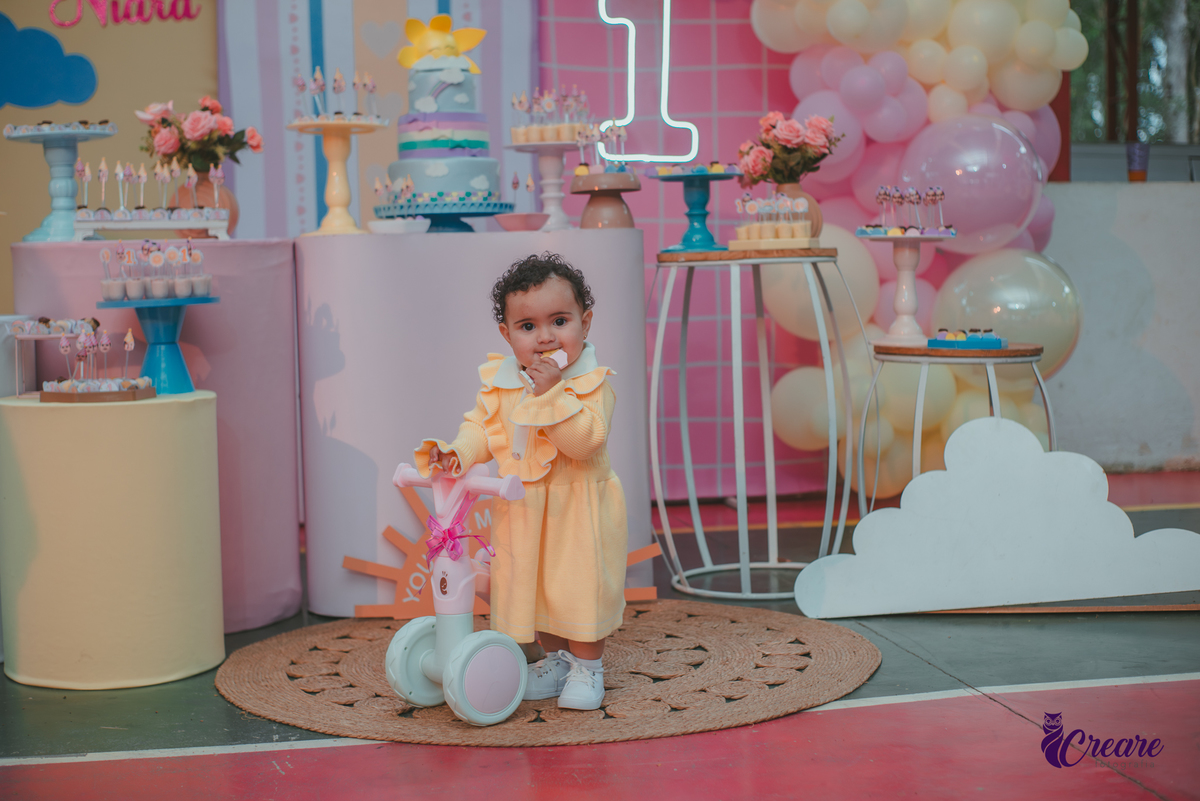 Uma bebê usando vestido amarelo e em frente mesa de decoração do seu aniversário de 1 ano.