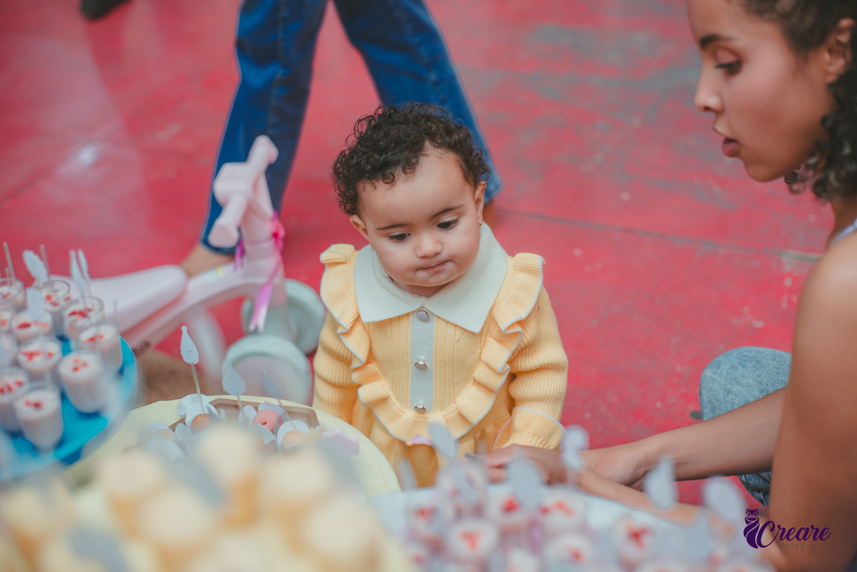 Uma bebê usando vestido amarelo e em frente mesa de decoração do seu aniversário de 1 ano.