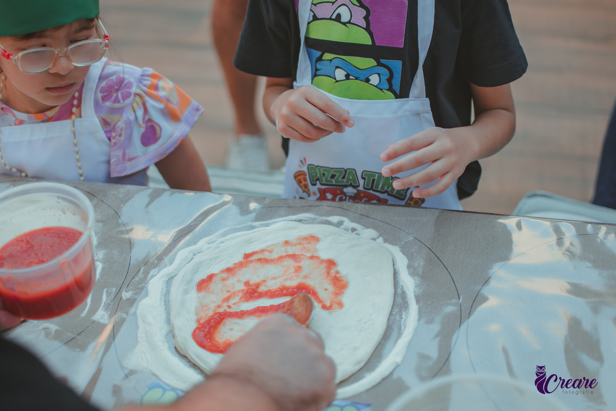 Crianças preparando pizza durante brincadeira de festa de aniversário infantil em casa.