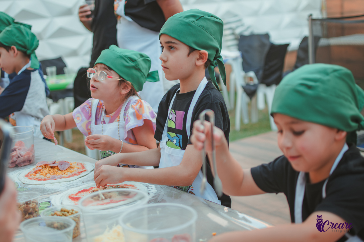 Crianças preparando pizza durante brincadeira de festa de aniversário infantil em casa.