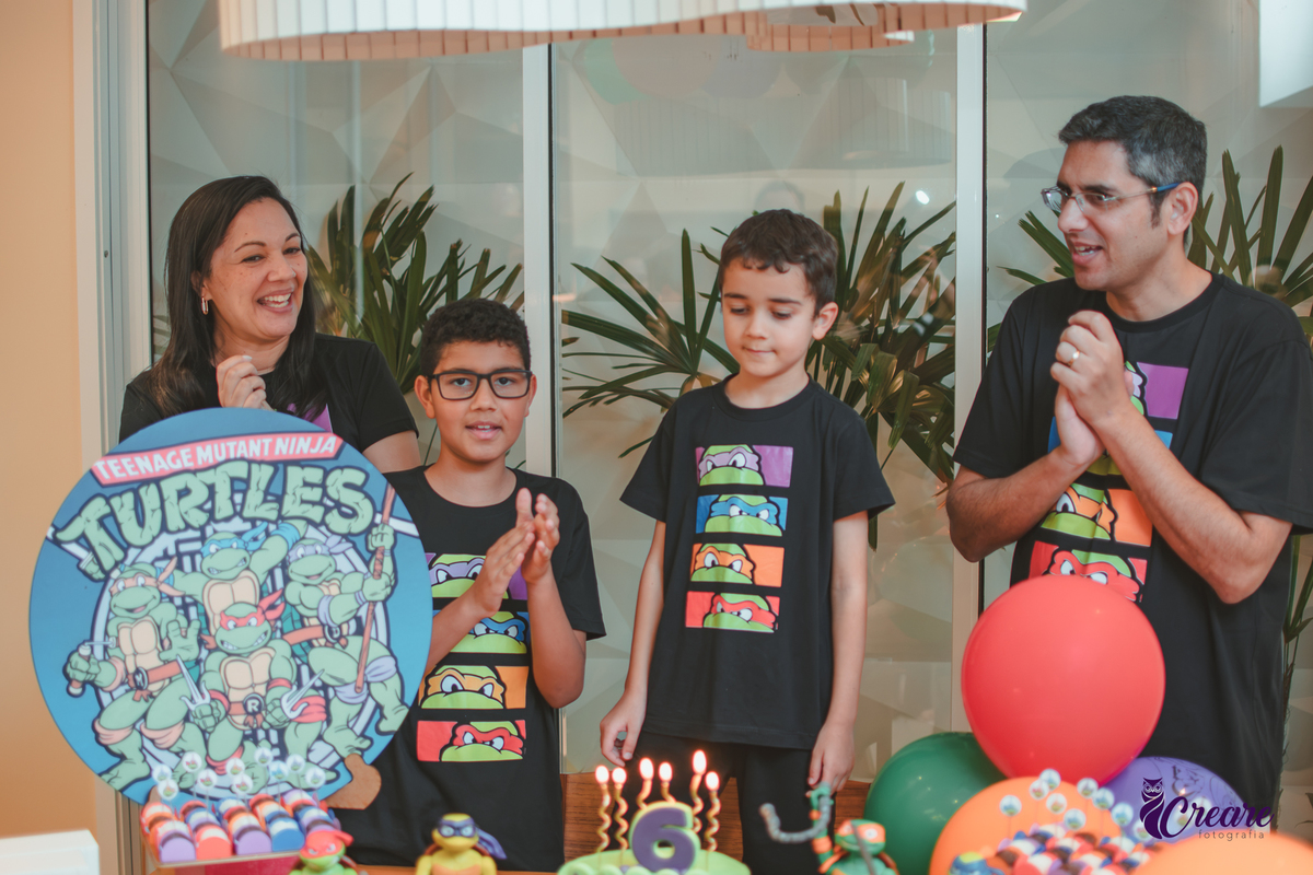 Uma família com pais, mãe e dois meninos, cantando parabéns para o menino de 6 anos, durante aniversário infantil.