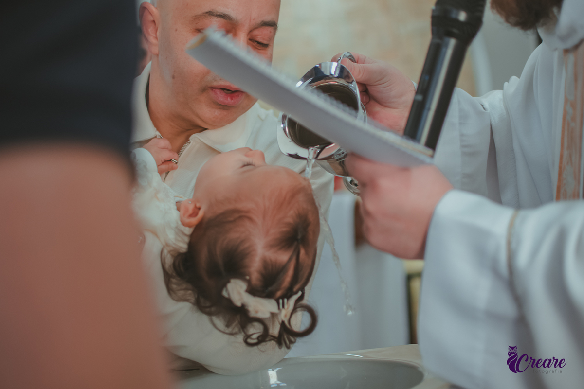 O Padre jogando água batismal na testa da bebê que está no colo de seu padrinho, durante batizado.