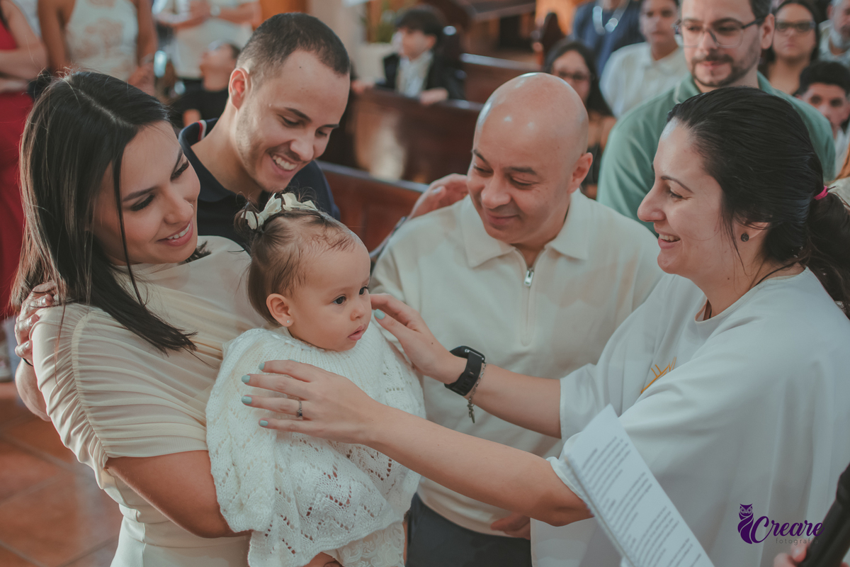 Uma mulher colocando o manto sagrado no bebê durante o batizado, em volta estão seu mãe que a segura, padrinho e pai.