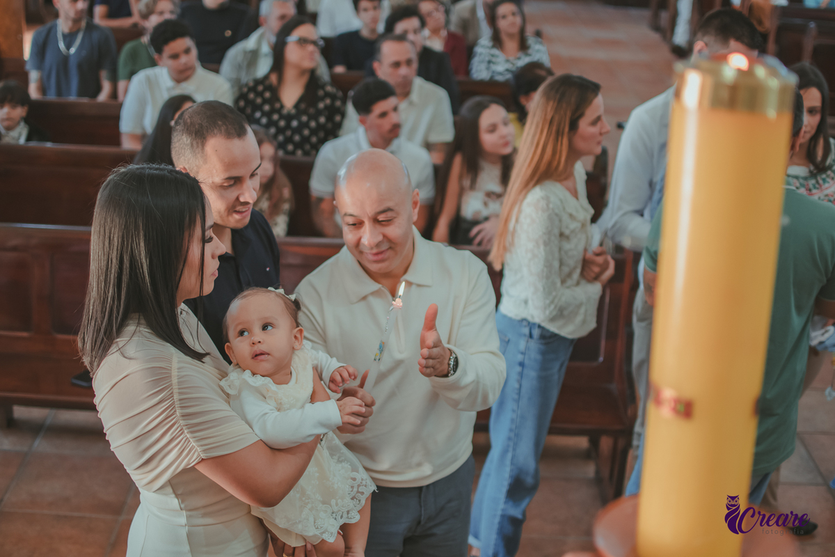 Um homem segurando uma vela de batizado ao lado do pai e mãe com o bebê no colo.