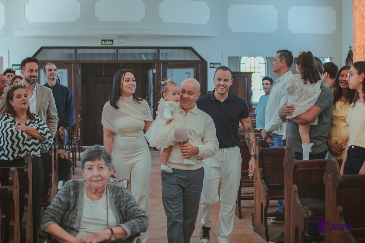 uma homem com  uma bebê no colo e uma casal o seguindo pelo corredor da igreja, na entrada para o batizado da criança.