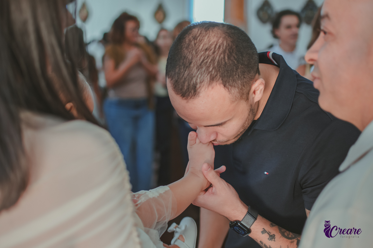 um homem beijando o pé de sua filha que está sem sapattos, durante batizado na igreja católica.