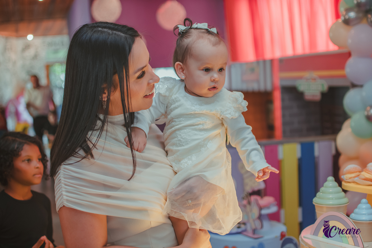 Uma mulher com seu bebê no colo, as duasolham a decoração da festa de 1 ano.