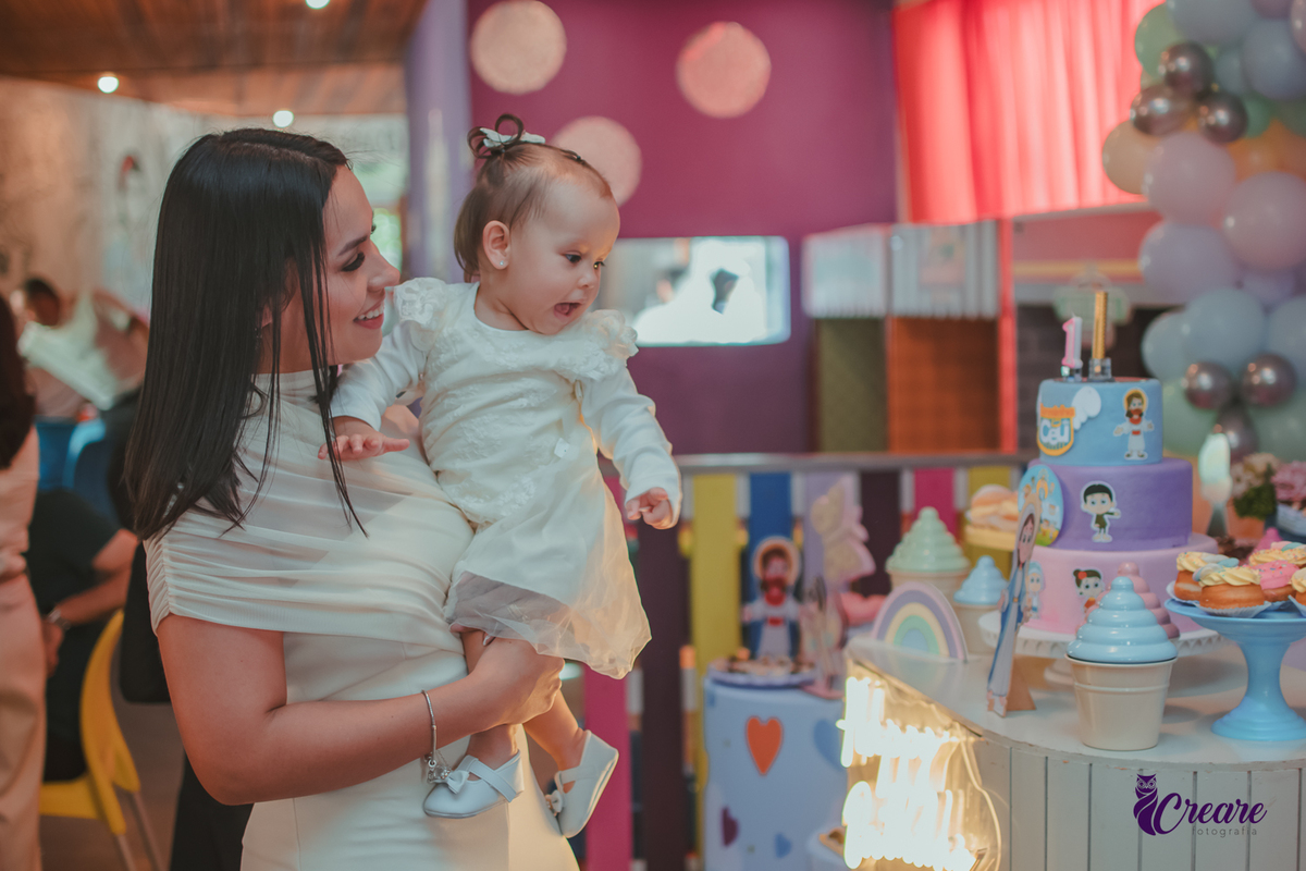 Uma mulher com seu bebê no colo, as duasolham a decoração da festa de 1 ano.