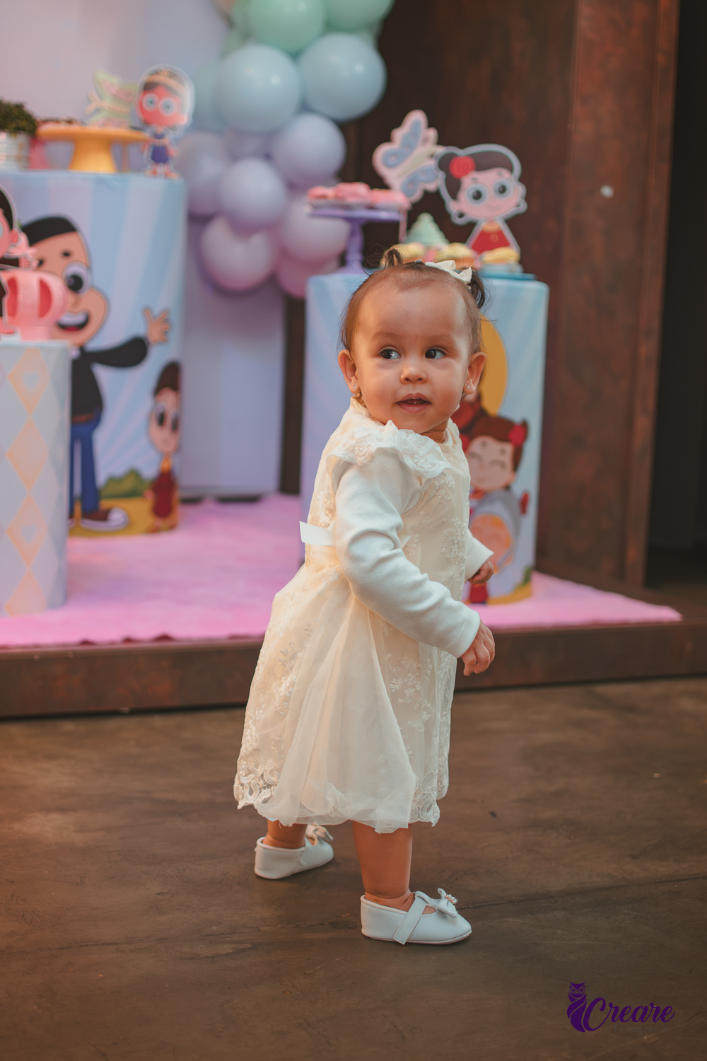 Uma bebê em pé e sorrindo na sua festa de aniversário de 1 ano.