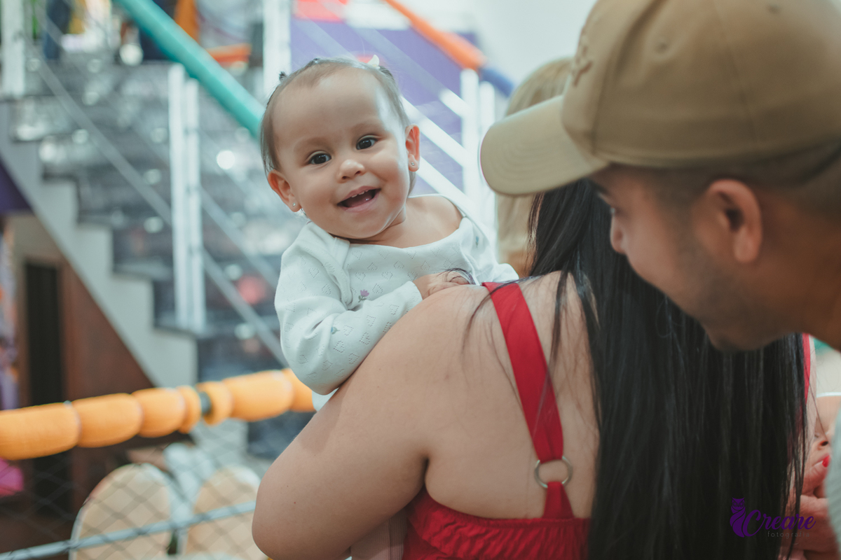 Uma bebê sorrindo no colo de uma mulher, enquanto um homem brinca com ela em sua festa de aniversário de 1 ano.