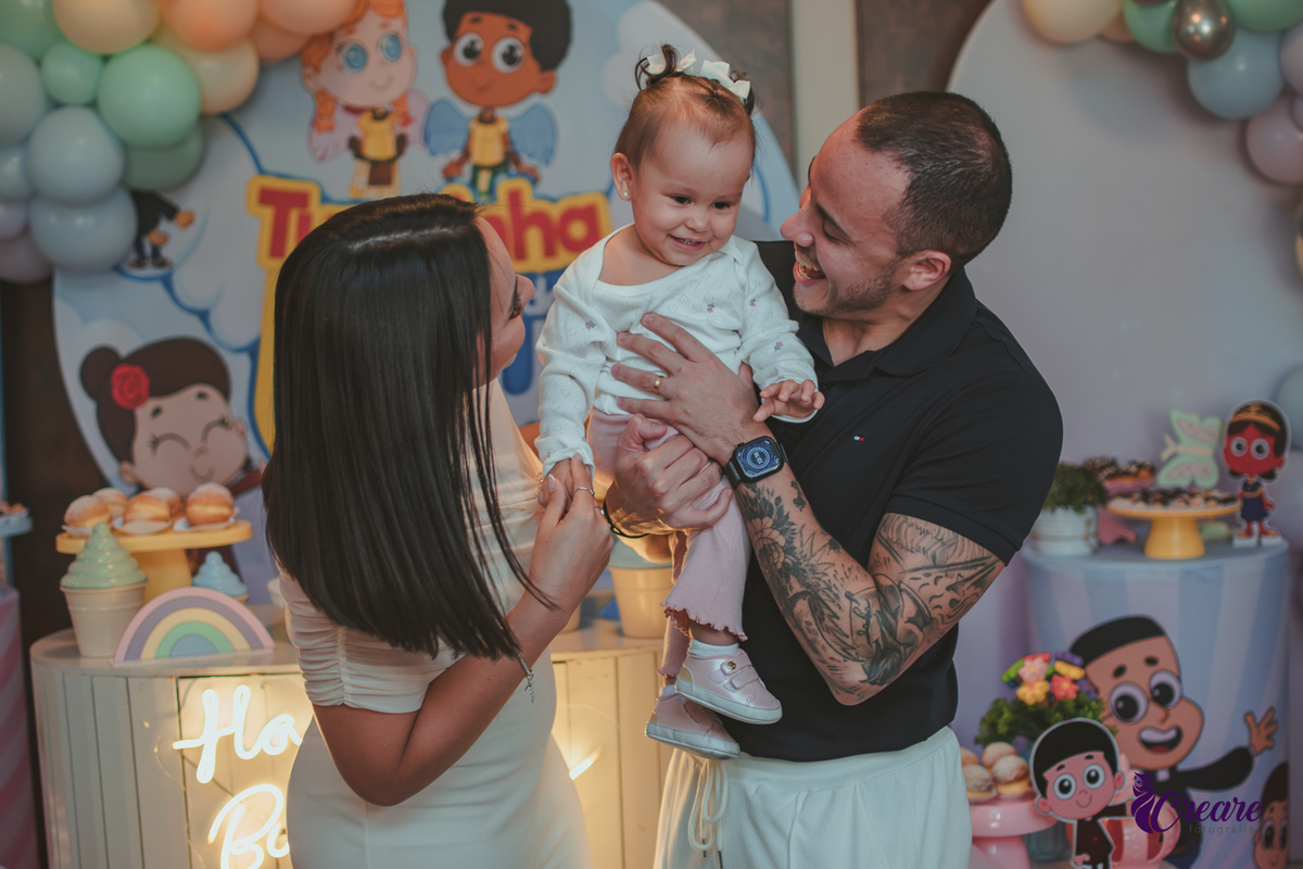 Uma familia  brincando com a bebê de 1 ano em frente a mesa de decoração da festa.